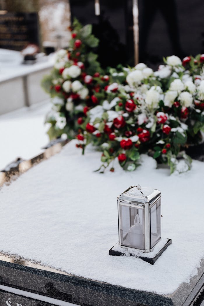 Snow-covered grave with flowers and a candle holder in a winter cemetery setting.