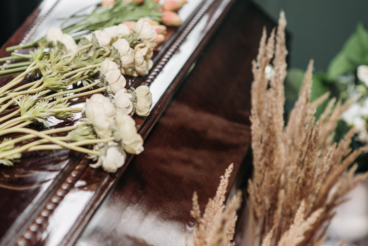A solemn funeral arrangement featuring a wooden casket adorned with white ranunculus and dried grasses.